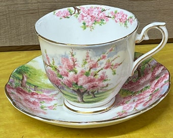 May include: White teacup and saucer set with a floral design featuring pink blossoms and a gold rim. The cup and saucer have a landscape scene with trees and a green field. The teacup has a curved handle.