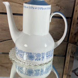 Może przedstawiać: Biały ceramiczny dzbanek do kawy z niebieskim wzorem kwiatowym i niebieską pokrywką. Dzbanek ma uchwyt i dzióbek.