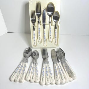 Puede incluir: Un juego de cubiertos vintage con cucharas, tenedores y cuchillos. Los mangos blancos tienen un diseño floral azul. El juego incluye un cuchillo, un tenedor y una cuchara, y está empaquetado en una bandeja de plástico.