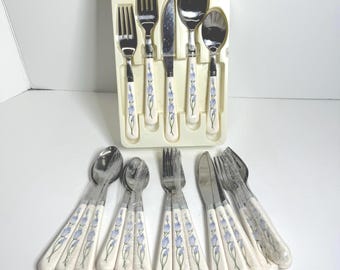 Op de afbeelding: Een vintage bestekset met lepels, vorken en messen. De witte handvatten hebben een blauw bloemmotief. De set bevat een mes, vork en lepel en is verpakt in een plastic bakje.