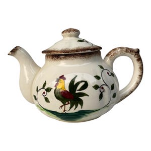 May include: Cream ceramic teapot with a rooster design. The teapot has a brown rim and handle, a matching lid, and a painted rooster with green, yellow, and red accents. Green vines and red berries decorate the sides.