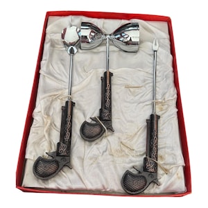Könnte beinhalten: Ein Vintage-Cocktail-Set in einer roten Box. Das Set enthält einen Löffel, einen bogenförmigen Sieb und eine Gabel, alle mit pistolenförmigen Griffen in Bronze- und Silbertönen. Das Set wird auf einem weißen, strukturierten Stoff präsentiert.
