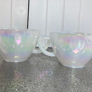 May include: Two white iridescent glass teacups with a diamond pattern. The cups have a handle and are made of opaque glass.