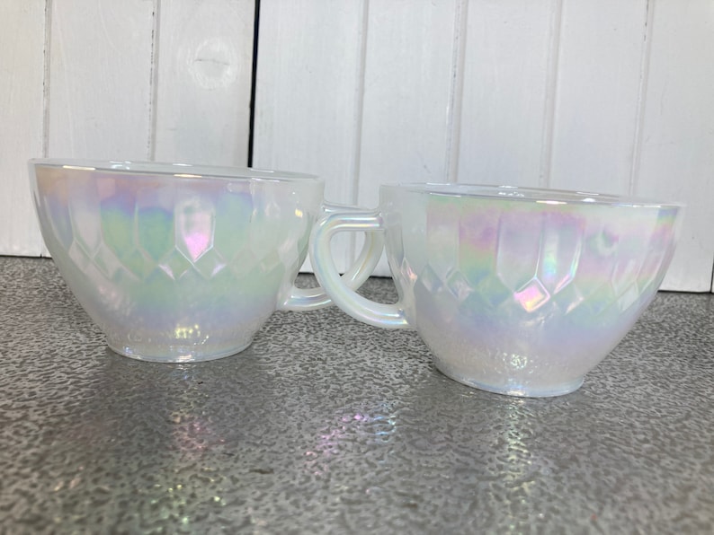 May include: Two white iridescent glass teacups with a diamond pattern. The cups have a handle and are made of opaque glass.