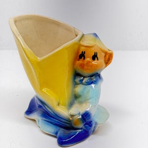 Op de afbeelding: Een keramische plantenpot in de vorm van een persoon die een gele en blauwe outfit draagt. De pot heeft een brede opening aan de bovenkant om te planten.