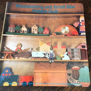 May include: A book titled "Woodworking and Scroll Saw Pattern Club" featuring a collection of large size patterns and craft ideas for all levels and skills of woodworking. The cover showcases a colourful illustration of a wooden shelf with various handcrafted items, including houses, a clock, a game board, a boat, sheep, a doll, a box, and a cactus.