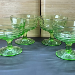 Puede incluir: Cuatro copas de cristal verde. Cada copa tiene un cuenco redondo con estrías horizontales y un tallo que se ensancha en una base circular. Las copas están dispuestas sobre una superficie oscura.
