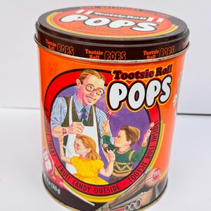 Könnte beinhalten: Eine zylindrische orangefarbene Tootsie Roll Pops-Dose mit braunem Deckel. Die Dose zeigt eine Vintage-Illustration eines Mannes und zweier Kinder, die Lutscher halten. Der Text enthält "Tootsie Roll Pops" und "Fruit Candy Outside, Tootsie Roll Inside!"