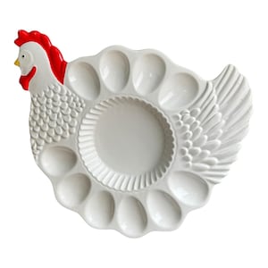 May include: White ceramic egg holder shaped like a hen. The hen features a red comb and wattle, a yellow beak, and a textured body. The egg holder has 12 egg-shaped indentations and a central circular area.