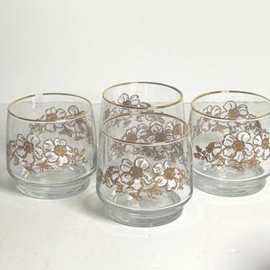 May include: Set of four clear glass tumblers with a gold rim and a white floral design. The flowers are outlined in gold.