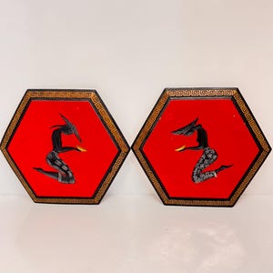 Puede incluir: Dos placas hexagonales de pared con fondo rojo y borde negro y dorado. Cada placa presenta una silueta negra de una mujer en posición de rodillas, sosteniendo un objeto dorado en su mano.
