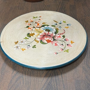 May include: A round, cream-colored ceramic plate with a hand-painted floral design. The flowers are pink, red, yellow, blue, and green. The plate has a teal blue rim.