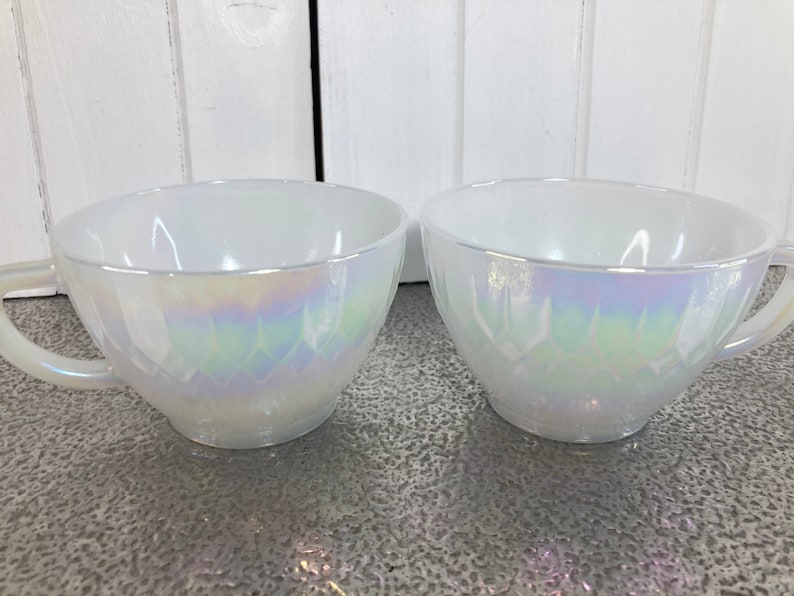 May include: Two white iridescent glass teacups with a diamond pattern. The cups have a handle on the left side.