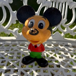 May include: A molded plastic figurine of a cartoon mouse character with large black ears, a tan face, a red shirt, green pants, and black shoes. The character has a small black nose with visible paint wear and wide white eyes.