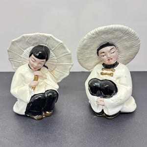 Peut inclure: Deux figurines en céramique représentant des personnages asiatiques. L'une tient un parasol blanc, l'autre porte un grand chapeau blanc. Les deux sont vêtus de vestes blanches, de pantalons noirs et d'accents dorés. Les figurines sont en position assise.