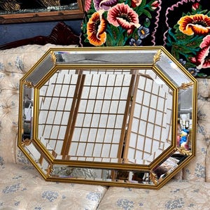 Op de afbeelding: Achtekantige spiegel met een gouden lijst en afgeschuinde randen. De spiegel reflecteert een kamer met een wit traliewerk.