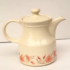Puede incluir: Tetera de cerámica color crema con tapa y asa. La tetera presenta un diseño floral en tonos coral y marrón alrededor de la base. El pico está en el lado derecho. La tetera es probablemente para servir té.