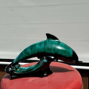 May include: A green and black ceramic dolphin figurine. The dolphin is leaping out of the water with its tail and fins extended. The figurine is glazed in a teal and black colour scheme.