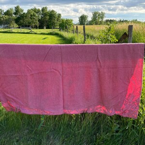 Op de afbeelding: Een roze stoffen laken hangt aan een houten hek tegen een groene grasveld met een huis en bomen op de achtergrond.