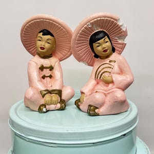 Peut inclure: Deux figurines asiatiques vintage. Chaque figurine est assise et porte une tenue rose et un grand chapeau rose en forme de soleil. Une figurine tient un éventail. Les figurines ont les cheveux noirs et sont assises sur une surface bleu clair.