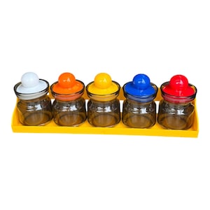 May include: Five glass spice jars with colorful lids in white, orange, yellow, blue, and red, arranged on a yellow tray. The jars are clear, allowing the contents to be visible. The lids have a rounded knob design.