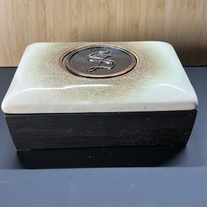 May include: A white ceramic box with a black wooden base. The top of the box has a circular design with a copper-colored letter "S" in the center.