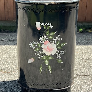 Könnte beinhalten: Schwarzer Wäschekorb aus Metall mit floralem Design mit rosa und weißen Rosen und grünen Blättern. Der Korb hat ein ausgeschnittenes Design am oberen und unteren Rand.