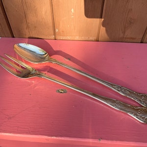 Puede incluir: Un juego de servicio de plata con un tenedor y una cuchara. Los utensilios tienen diseños ornamentados y están sobre una superficie rosa.