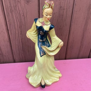 Puede incluir: Una figurita de cerámica de una mujer con un vestido amarillo y azul con un diseño dorado. Lleva un tocado con un diseño floral. La figurita está de pie sobre una superficie rosa.