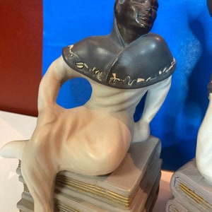 May include: A ceramic figurine of a seated figure with dark skin, wearing a dark gray cowl and light-colored clothing. The figure is perched atop a stack of gray books with gold-colored edges. The background is blue.