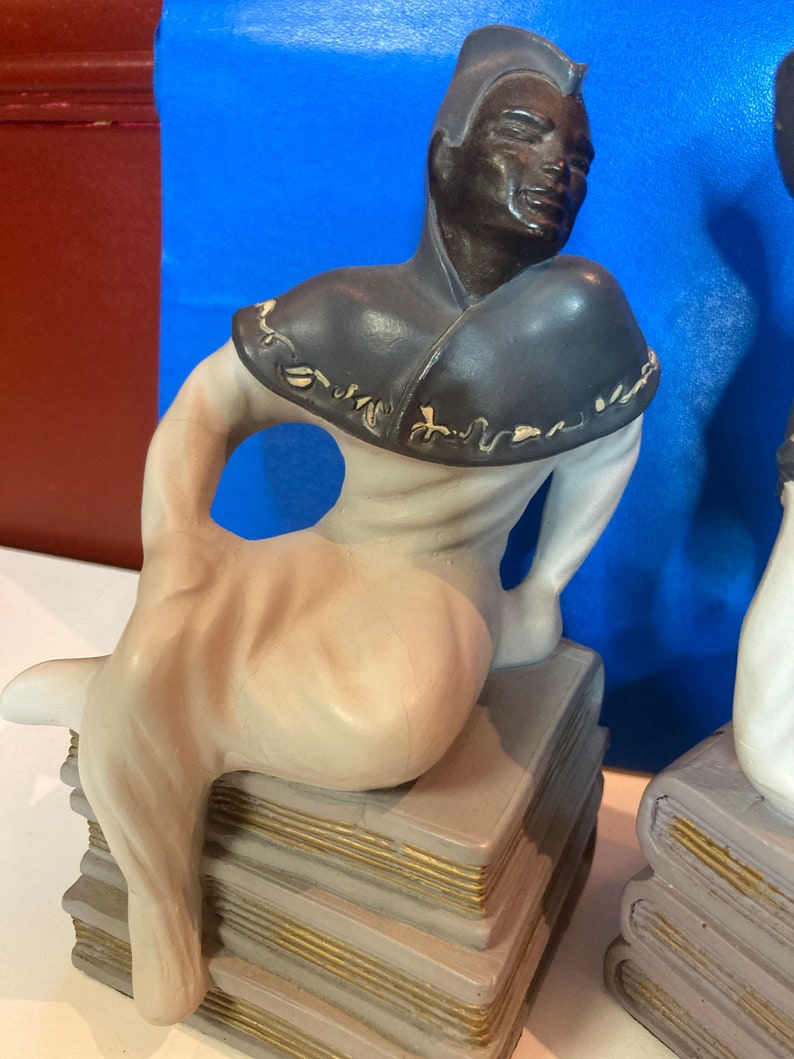 May include: A ceramic figurine of a seated figure with dark skin, wearing a dark gray cowl and light-colored clothing. The figure is perched atop a stack of gray books with gold-colored edges. The background is blue.