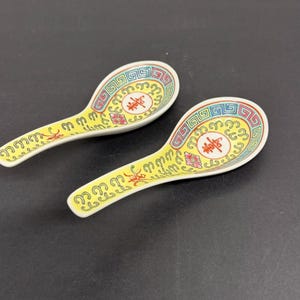 May include: Two yellow ceramic soup spoons with a decorative design. The spoons have a white rim with a red, green, and blue border. The spoon bowls feature a red symbol and floral accents.