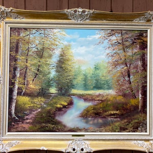 Puede incluir: Una pintura al óleo enmarcada que representa una escena de bosque con un arroyo sinuoso en primer plano. Los árboles son una mezcla de verde y marrón, y el cielo es azul claro con nubes blancas. La pintura está en un marco dorado con detalles ornamentados.