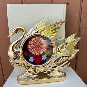 Peut inclure: Une horloge dorée en forme de cygne avec un cadran rond orné de chiffres rouges et dorés et de motifs floraux. L'horloge comporte deux cygnes aux ailes déployées et une base décorative. L'horloge est probablement vintage ou rétro.
