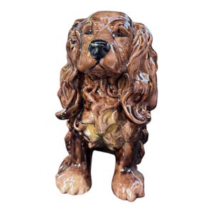 May include: A ceramic dog figurine in shades of brown, with a glossy finish. The dog has long, curly fur, a black nose, and a gentle expression. The figurine is standing, with its front paws slightly forward.