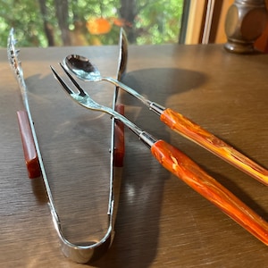 May include: A set of silver serving utensils with orange and yellow marbled handles. The set includes a fork, a spoon, and a pair of tongs.