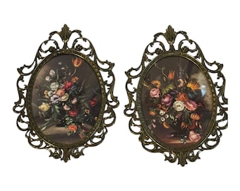 May include: Two vintage oval wall art pieces. Each features a floral still life painting in a dark oval frame with ornate, openwork detailing. The frames have a bronze finish, and the paintings display a variety of colorful flowers.