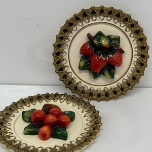 May include: Two decorative wall plates with a white background and gold trim. One plate features a cluster of red strawberries with green leaves. The other plate features a cluster of red cherries with green leaves.