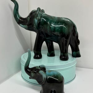 May include: Two ceramic elephant figurines. The larger elephant is dark green with a glossy finish, standing on a light blue tin. The smaller elephant is in front, with a similar color scheme. Both have raised trunks.
