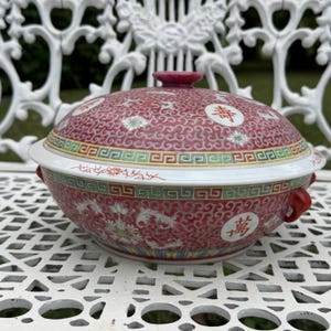 May include: A round ceramic lidded tureen featuring a vibrant red background with intricate white scrollwork patterns. The bowl and lid are decorated with colorful floral motifs and circular medallions containing red Chinese characters. It has two red handles.