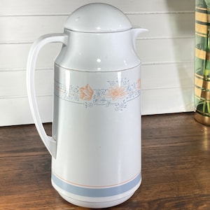 May include: White ceramic thermal carafe with a floral design in blue and orange. The carafe has a lid and a handle.