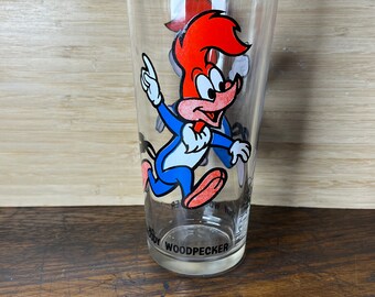 May include: A clear glass featuring a cartoon image of Woody Woodpecker running. The glass has "Woody Woodpecker" printed on the base.