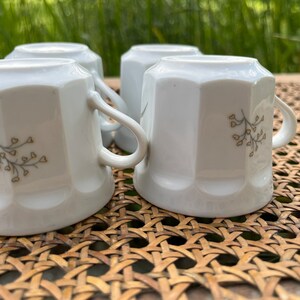 May include: Four white ceramic mugs with a delicate floral design in brown. The mugs are sitting on a woven wicker surface.