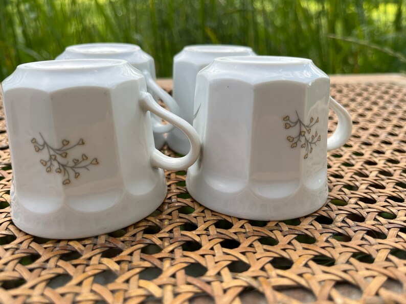 May include: Four white ceramic mugs with a delicate floral design in brown. The mugs are sitting on a woven wicker surface.