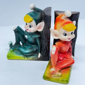 Peut inclure: Une paire de serre-livres en céramique en forme d'elfes. L'elfe de gauche porte une tenue verte et est assis sur une base verte. L'elfe de droite porte une tenue orange et est assis sur une base verte. Les deux elfes sont assis sur un tronc d'arbre brun.