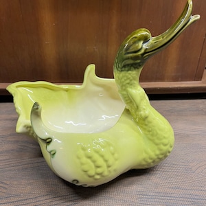 May include: A green ceramic planter shaped like a swan with a raised head and an open beak. The planter has a textured surface and a white interior.