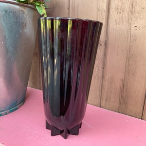 Könnte beinhalten: Eine hohe, dunkelburgunderfarbene Vase mit geriffeltem Design. Die Vase hat einen sternförmigen Sockel und eine ausgestellte Oberseite. Die Vase besteht aus glänzendem Material und steht vor einem Holzgrund.