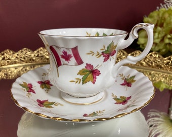 May include: A white teacup and saucer with a gold trim. The teacup features a red and white Canadian flag and a floral design with green and red leaves.