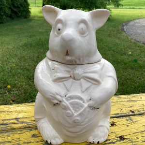 May include: White ceramic cookie jar shaped like a pig wearing a bow tie. The pig has a surprised expression and is holding a round object in its front paws.