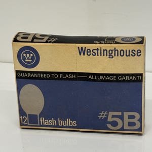 Puede incluir: Una caja marrón con una etiqueta azul y blanca que contiene 12 bombillas flash. La etiqueta dice "Westinghouse" y "#5B bombillas flash".